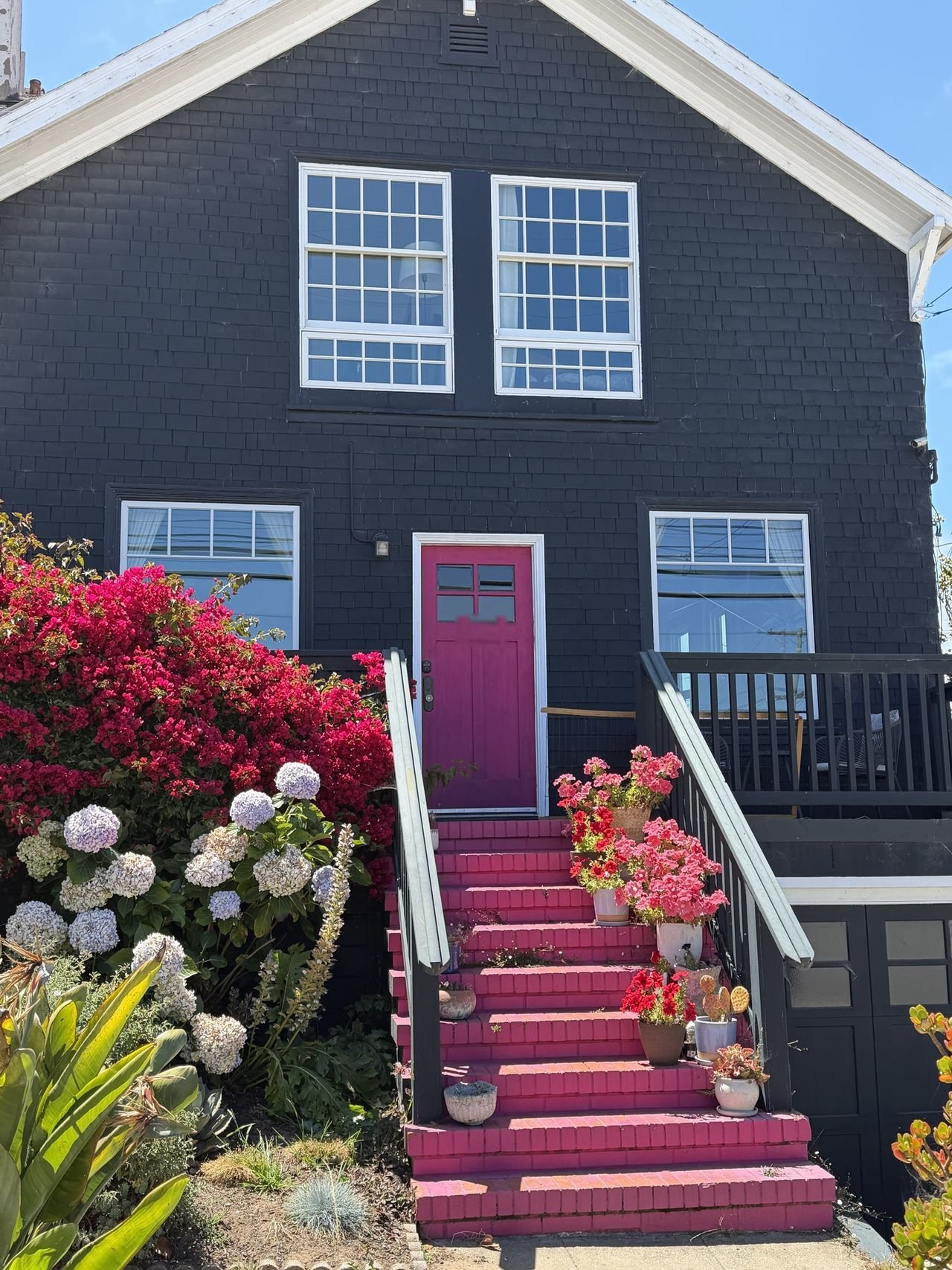 A dark shingled home with white-trimmed windows, a pink door and flower-lined pink steps