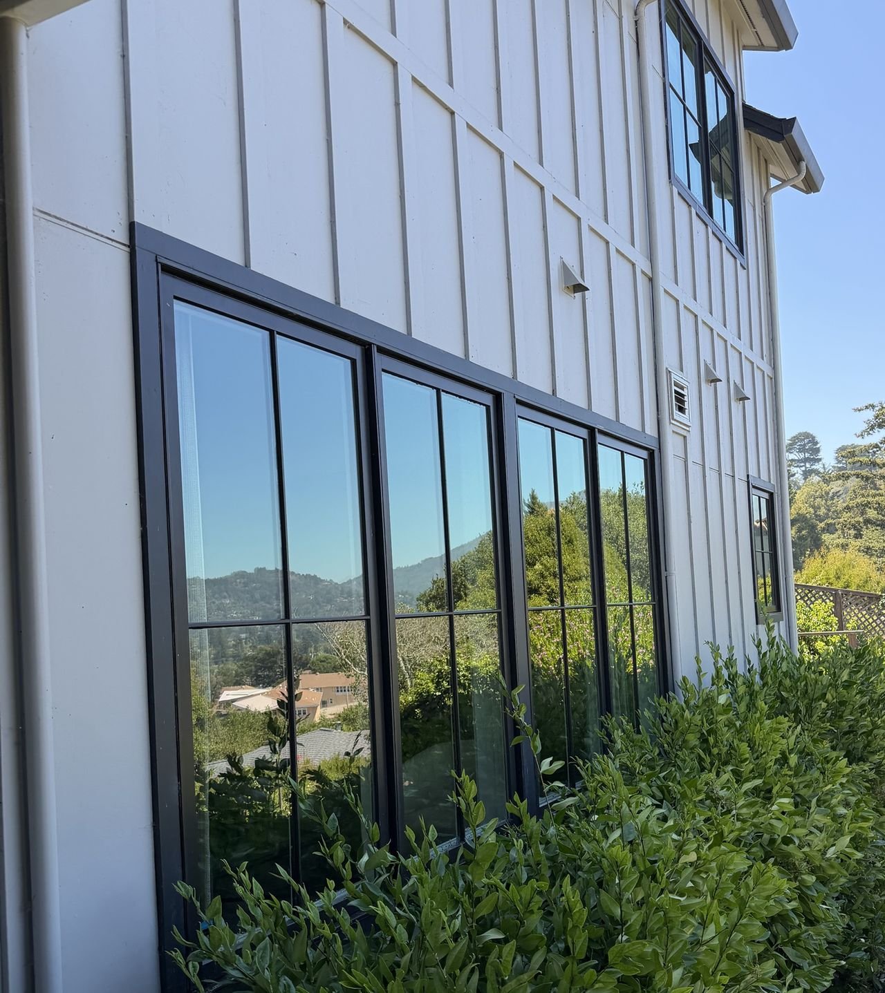 Clean black-framed windows on a white board-and-batten home in Mill Valley, reflecting trees, rooftops and the hills