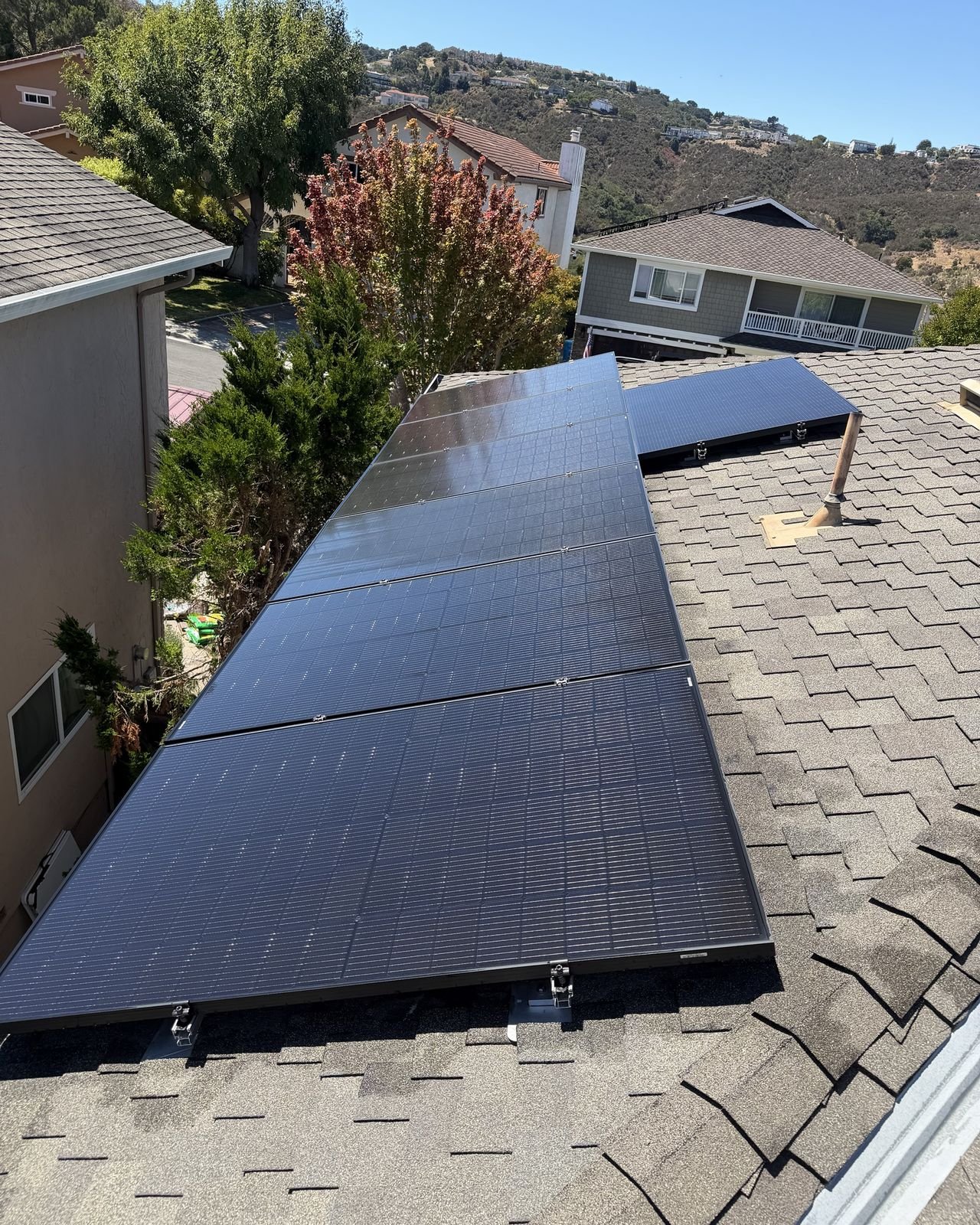 A row of freshly cleaned black solar panels on a shingled roof, with green hills and homes behind