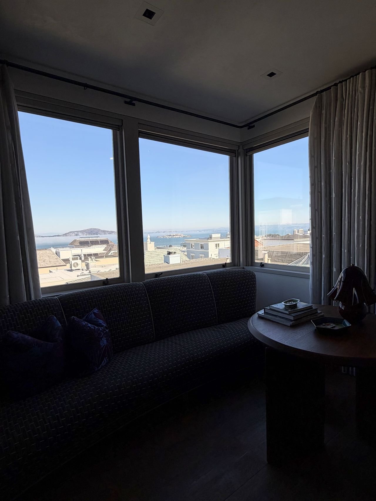 A dim living room with a curved sofa facing three clean corner windows that look out over rooftops to Alcatraz, Angel Island and the bay